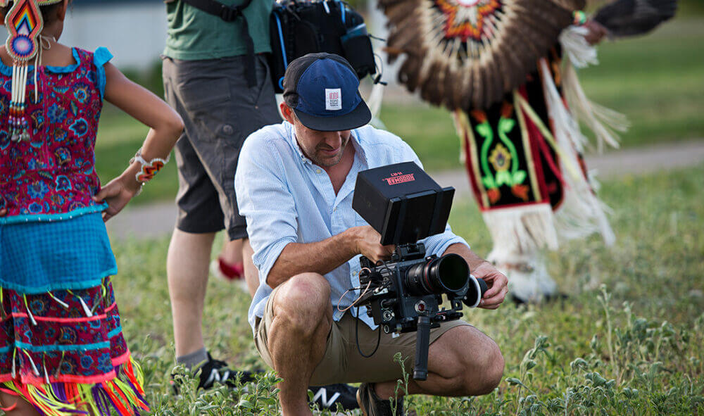 A videographyer preparing to film, with American Indian dancers in the background